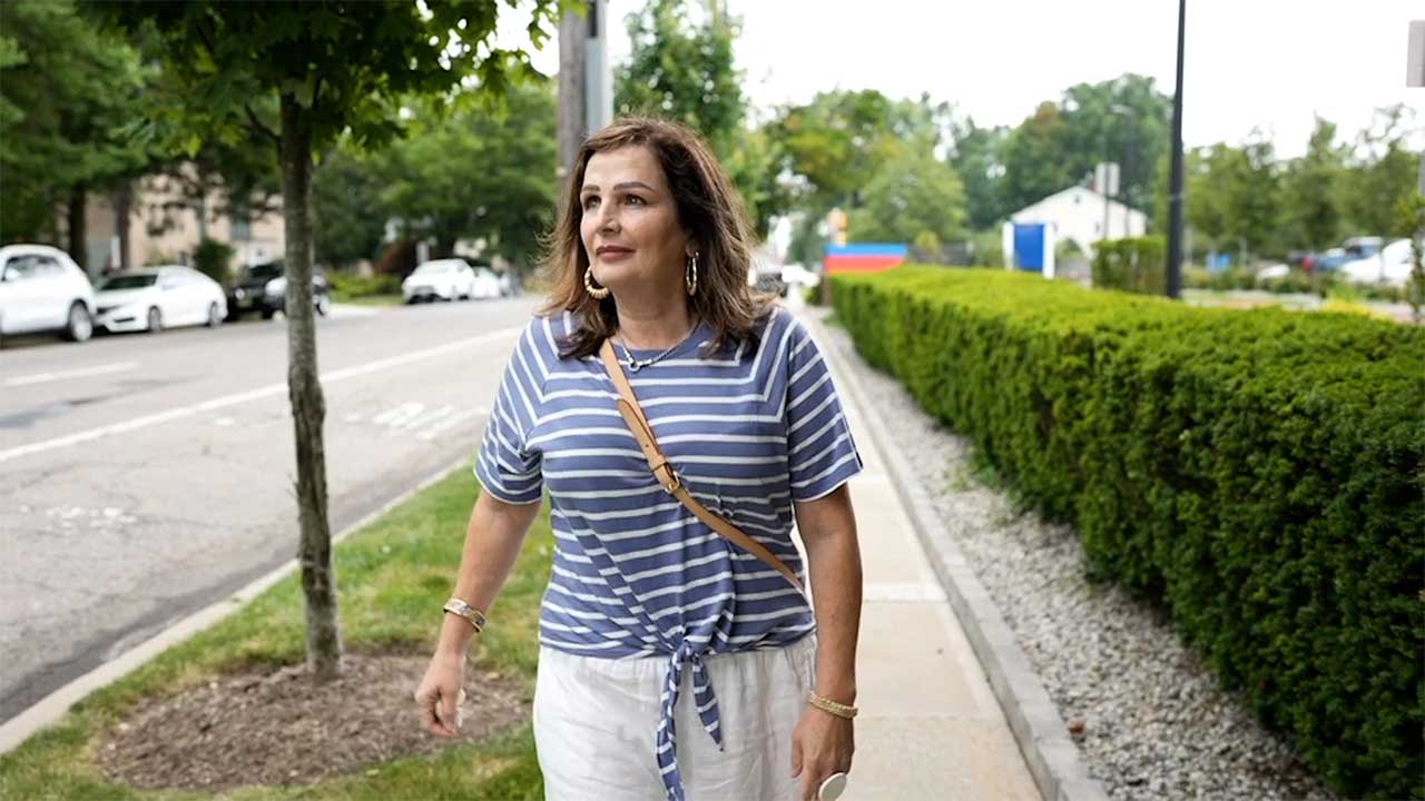 woman walking down a side walk with a white and blue strip shirt, white pants, and carrying an over the shoulder bag